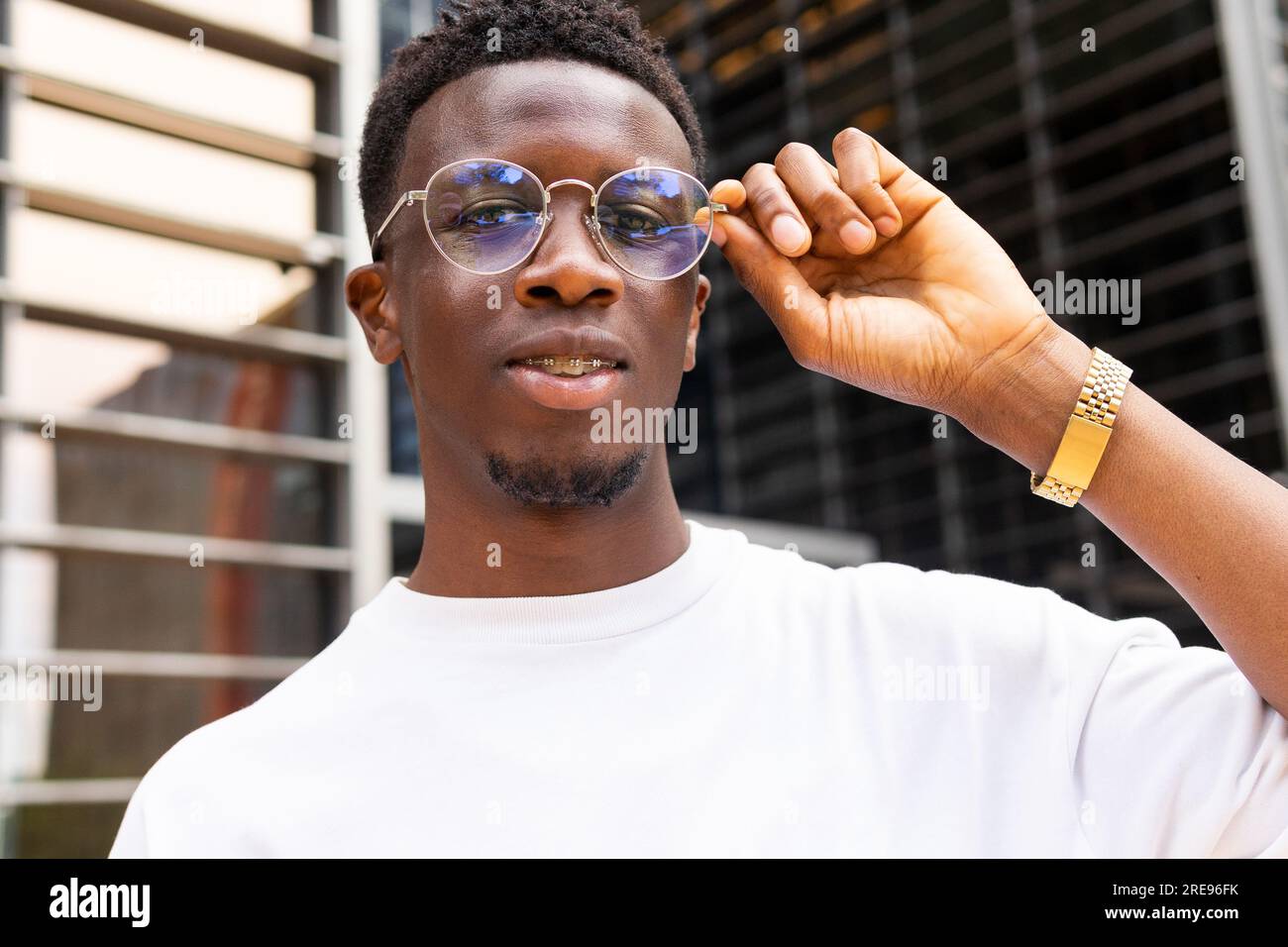 Ein seriöser afroamerikanischer Mann in trendigen Outfits und einer Brille, der vor modernen Gebäuden in die Kamera schaut Stockfoto