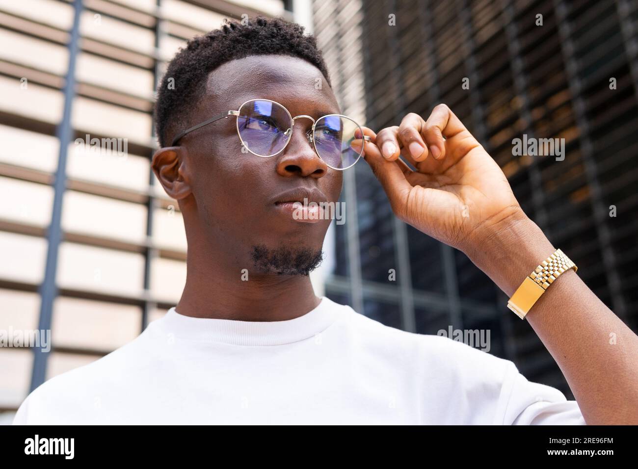 Ein seriöser afroamerikanischer Mann in trendigen Outfits und einer Brille, der wegsieht, während er sich gegen moderne Gebäude stellt Stockfoto