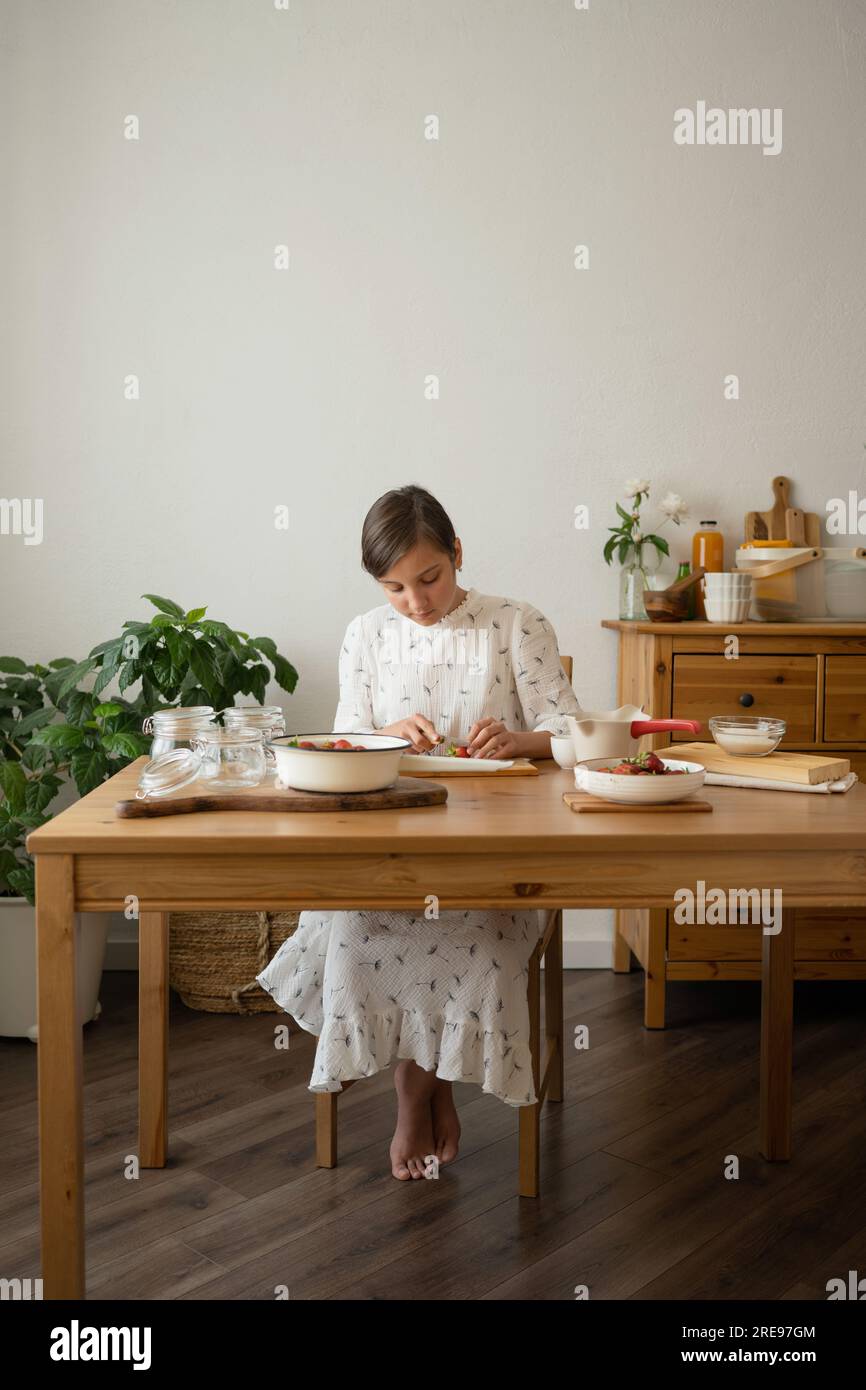 Der ganze Körper einer jungen Frau im Kleid sitzt am Holztisch mit einer Schüssel Erdbeeren und Glasflaschen, während sie Erdbeermarmelade im Zimmer macht Stockfoto