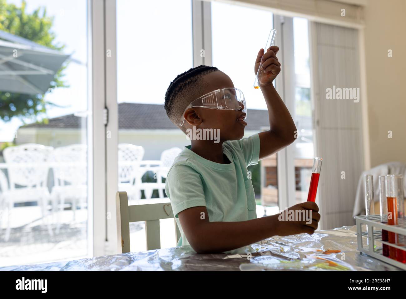 Fokussierter afroamerikanischer Junge, der ein Chemie-Experiment macht und sich ein Reagenzglas im sonnigen Wohnzimmer anschaut Stockfoto