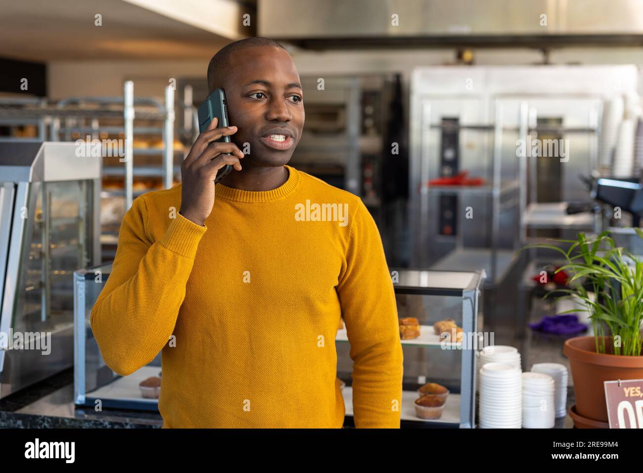 Glücklicher afroamerikanischer Bäckereiangestellter, der ein gelbes Sweatshirt trägt und mit dem Smartphone spricht Stockfoto