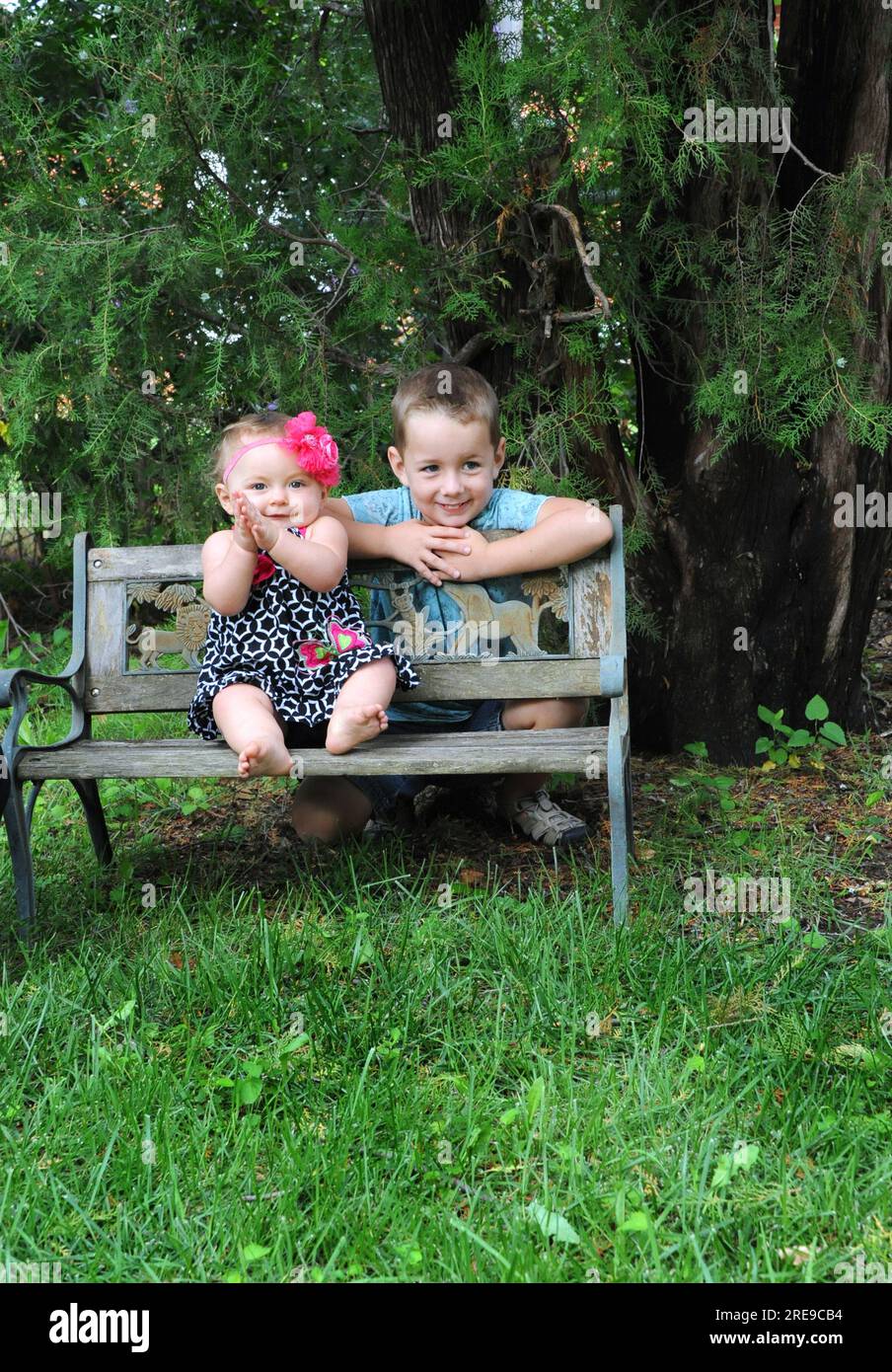 Schwester und Bruder stehen Seite an Seite. Der Bruder lehnt sich über die Parkbank und die Schwester sitzt auf dem Sitz. Stockfoto