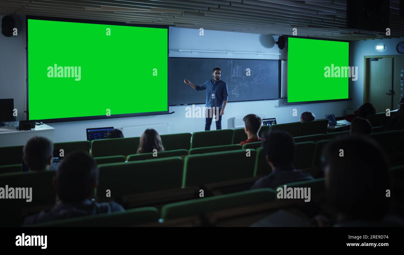 Junger Universitätsprofessor erklärt einen Vortrag, zeigt auf ein Mockup-Display mit grünem Bildschirm. Eine Gruppe von multiethnischen Schülern Stockfoto