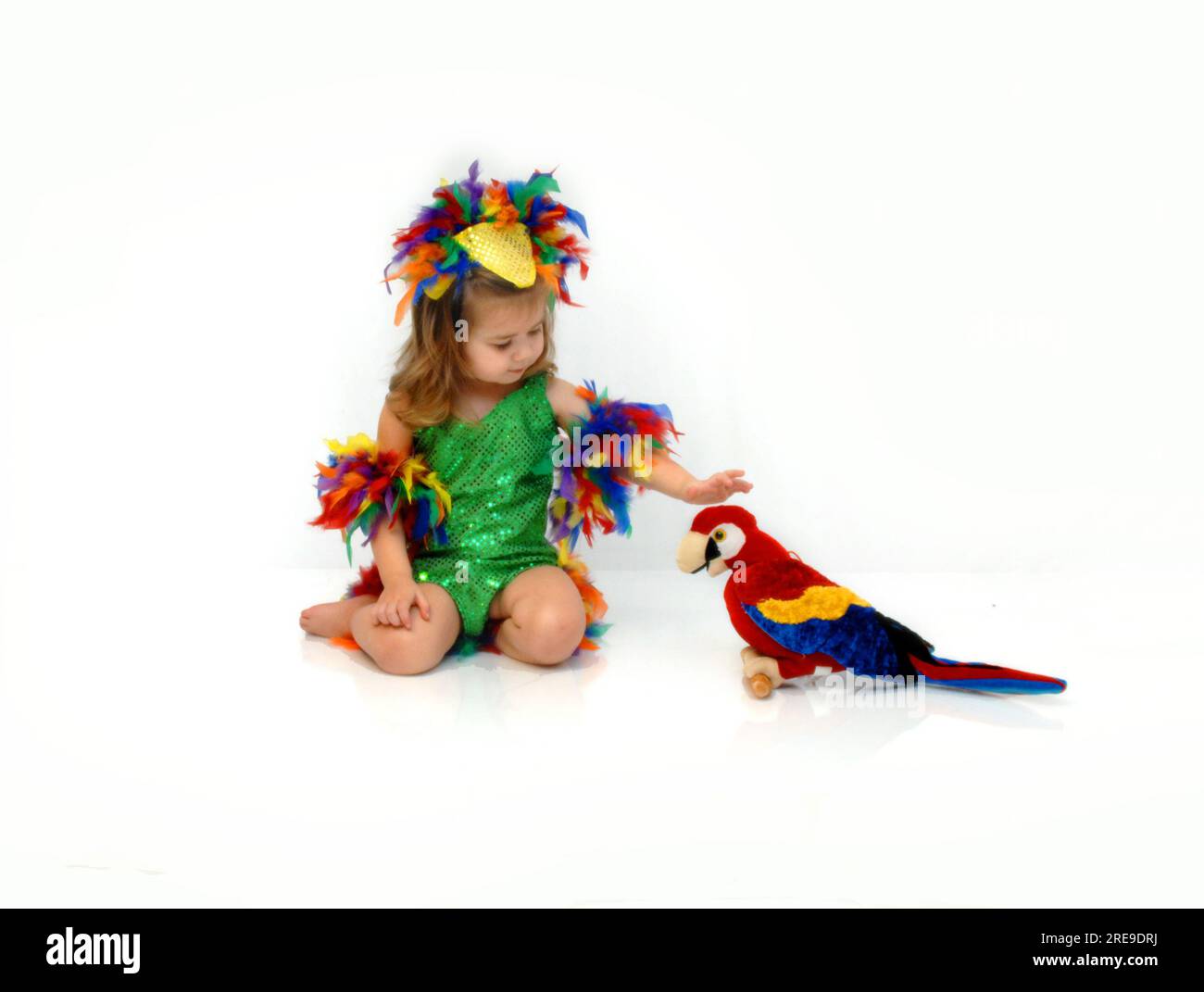 Das niedliche kleine Mädchen trägt ein farbenfrohes Federkostüm mit leuchtend grünen Pailletten. Sie streichelt ihren Papagei. Stockfoto