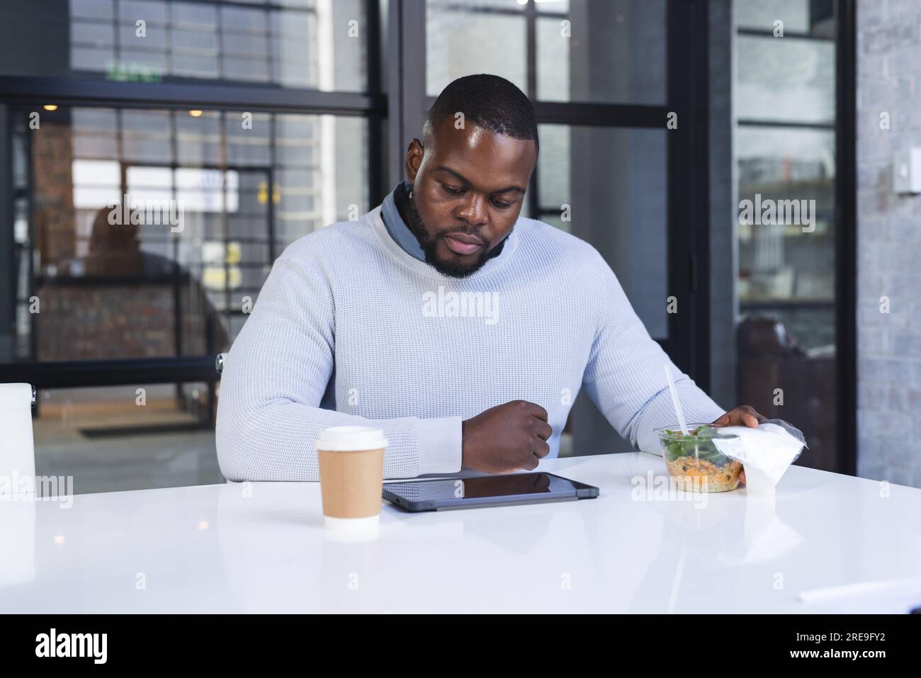 Fokussierter, ungezwungener afroamerikanischer Geschäftsmann, der mit Kaffee zu Mittag isst und ein Tablet benutzt Stockfoto