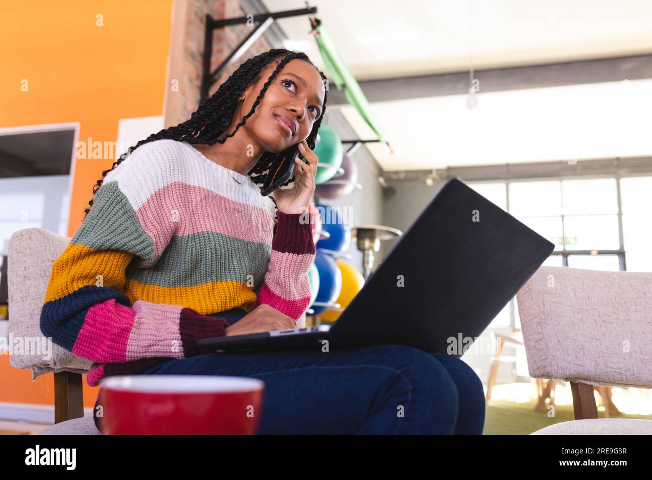 Eine afroamerikanische Geschäftsfrau, die im Büro mit einem Smartphone spricht und ein Notebook verwendet Stockfoto