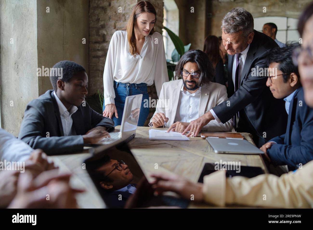 Ein multikulturelles Business-Team versammelt sich um einen Tisch, Laptops stehen zur Verfügung. Ein junger Unternehmer aus dem Nahen Osten unterzeichnet Dokumente, die von einem Senior in s geleitet werden Stockfoto