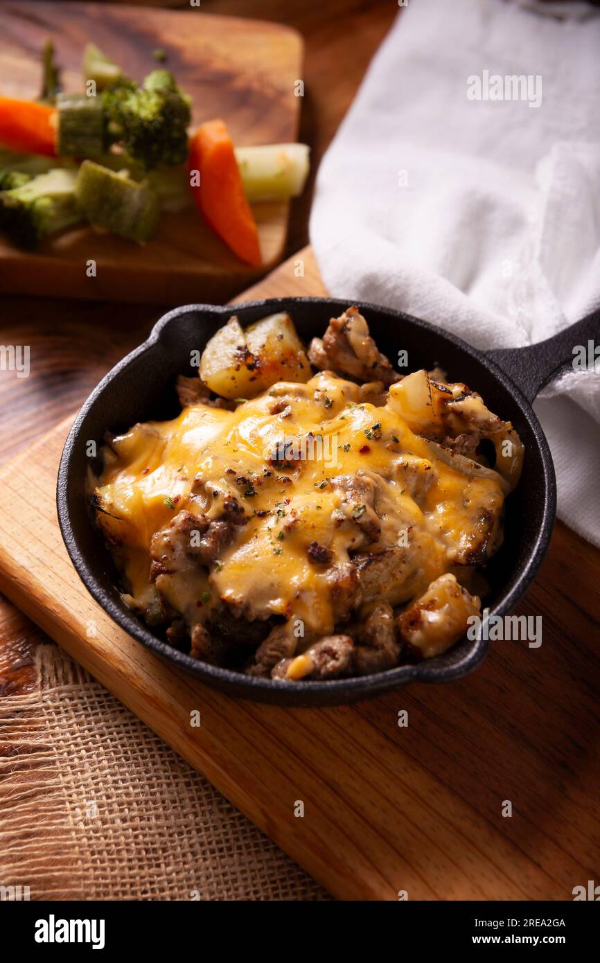 Hausgemachtes Rezept für Kartoffeln mit Steak und Schmelzkäse, gewürzt und serviert in einer gusseisernen Pfanne auf einem rustikalen Holztisch. Stockfoto