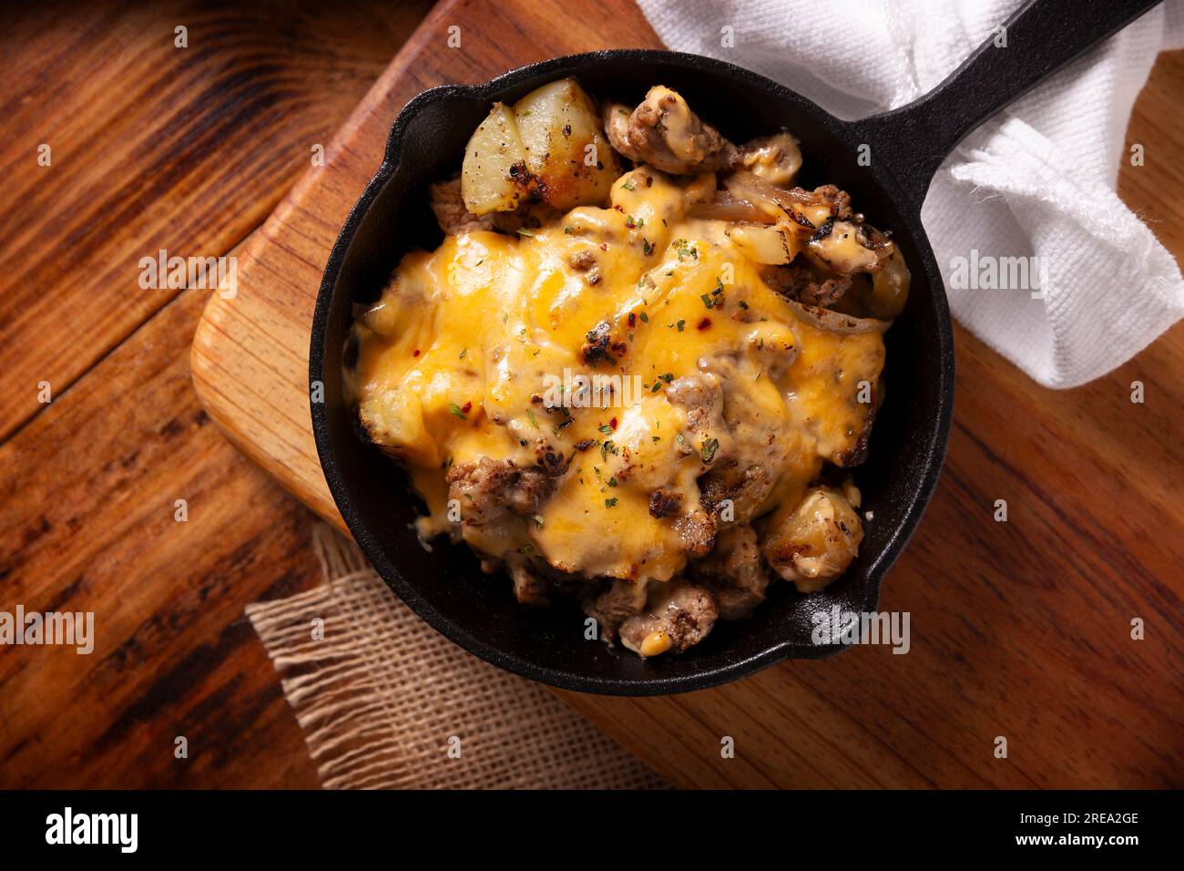 Hausgemachtes Rezept für Kartoffeln mit Steak und Schmelzkäse, gewürzt und serviert in einer gusseisernen Pfanne auf einem rustikalen Holztisch. Tabelle topview. Stockfoto