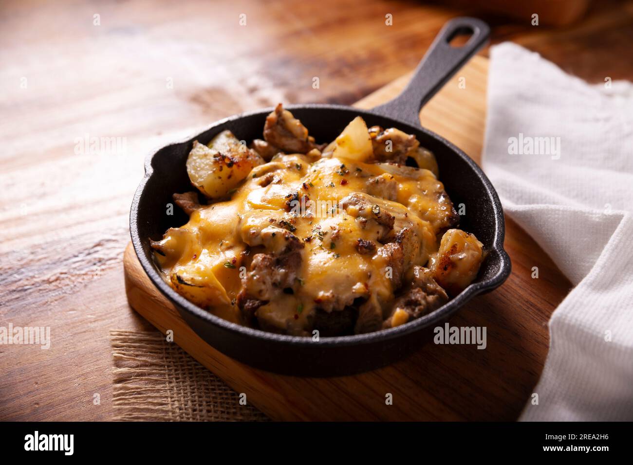 Hausgemachtes Rezept für Kartoffeln mit Steak und Schmelzkäse, gewürzt und serviert in einer gusseisernen Pfanne auf einem rustikalen Holztisch. Stockfoto