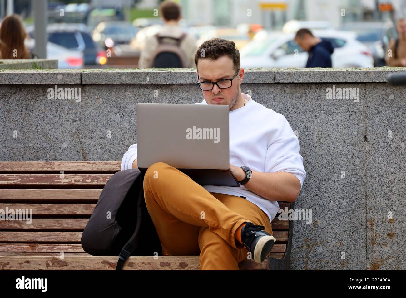 Ein Typ mit Brille, der mit einem Laptop auf einer Bank auf der Sommerstraße sitzt Stockfoto
