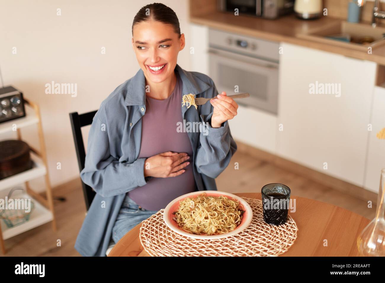 Zufriedene junge schwangere Frau, die hausgemachte Pasta probiert, am Tisch in der Küche sitzt, Kopierraum Stockfoto