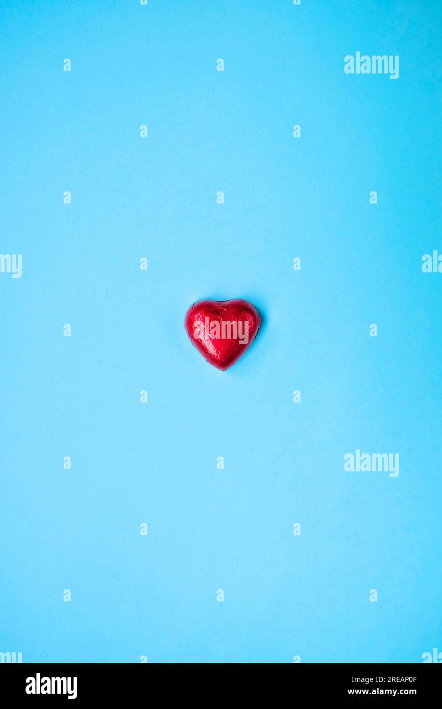Ein kleines rotes Herz auf blauem Hintergrund, eine Schokolade in Alufolie verpackt. Stockfoto