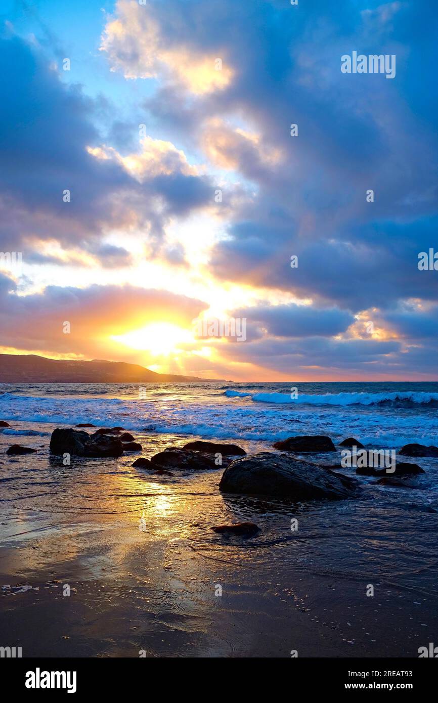 Dunkle nasse große Steine am Strand am Wasserrand bei Sonnenuntergang. Meereslandschaft mit schweren dunklen Wolken und untergehender Sonne. Stockfoto