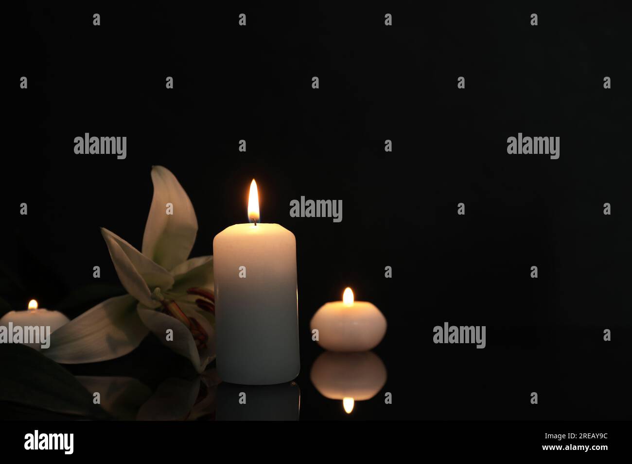 Weiße Lilie und brennende Kerzen auf schwarzer Spiegeloberfläche bei Dunkelheit, Platz für Text. Begräbnissymbole Stockfoto