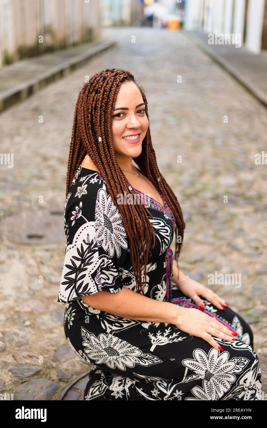 Eine Frau hockte auf einer Kopfsteinpflasterstraße. Haargeflechte und afrikanisches Kleid. Pelourinho, Brasilien. Stockfoto