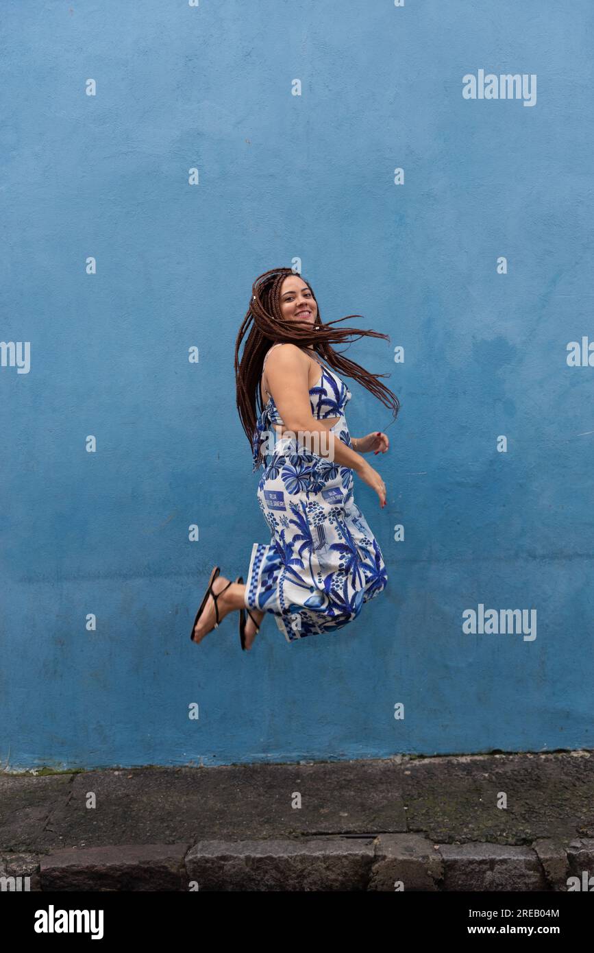 Porträt einer schönen Frau, die mit Zöpfen im Haar gegen die blaue Wand springt. Pelourinho, Brasilien. Stockfoto