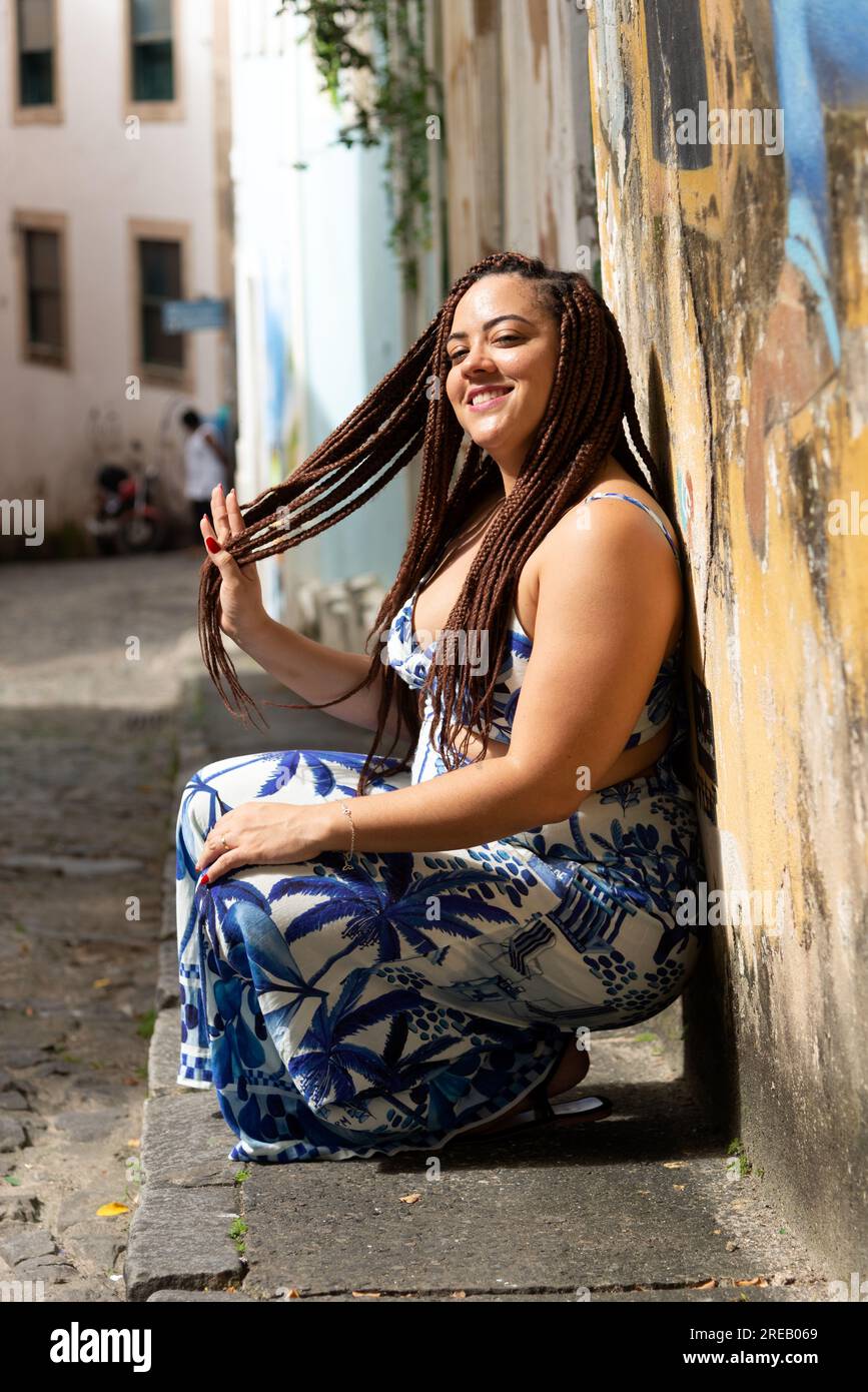 Schöne Frau mit geflochtenen Haaren, die sich an eine Wand lehnt, mit Straßen im Hintergrund. Pelourinho, Brasilien. Stockfoto