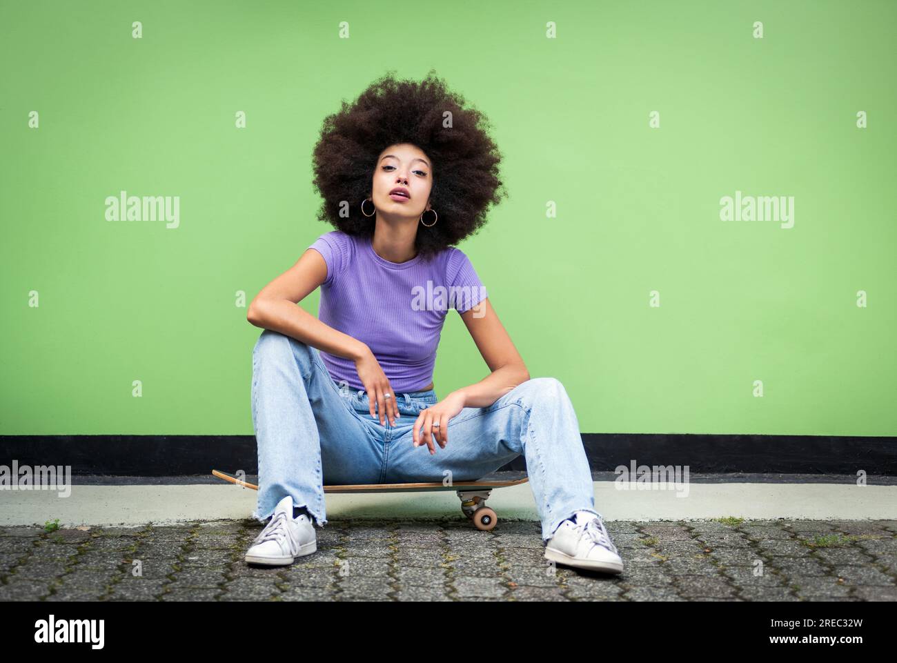 Der ganze Körper einer jungen afrikanischen Frau in Afro-Frisur und legerer Kleidung mit Make-up sitzt auf dem Skateboard über dem Terrassenboden und schaut auf Kamera aga Stockfoto