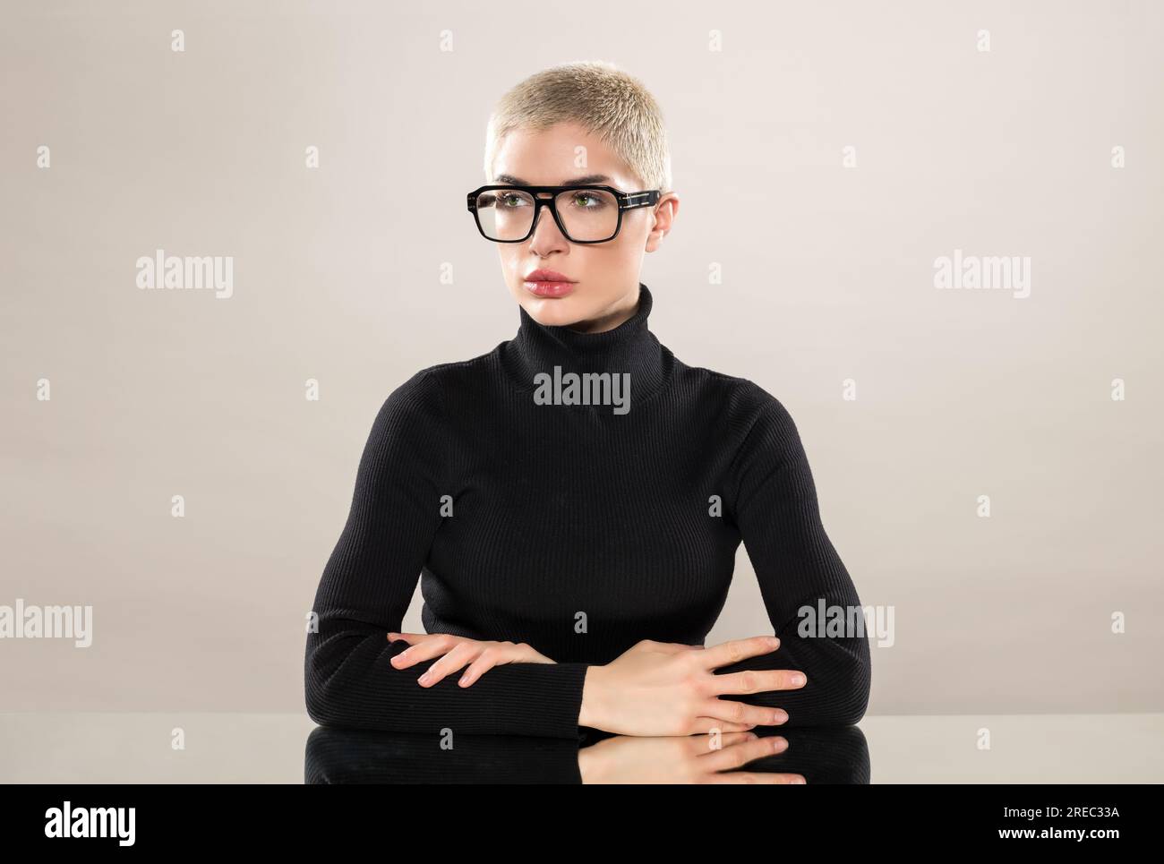 Seriöses weibliches Modell mit schwarzem Rollkragenpullover und Brille kreuzt die Arme, während es auf einem Spiegeltisch auf grauem Hintergrund sitzt und wegschaut Stockfoto