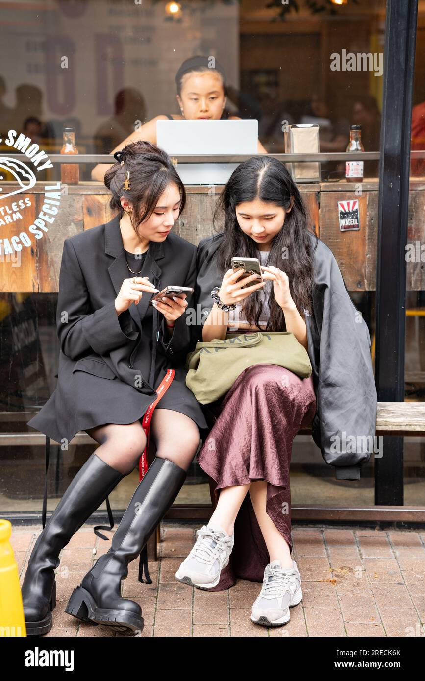 Zwei asiatische Schülerinnen, die auf iphones bei der Royal College of Art Summer Show in Truman Gallery 2023 SMS schreiben. Stockfoto