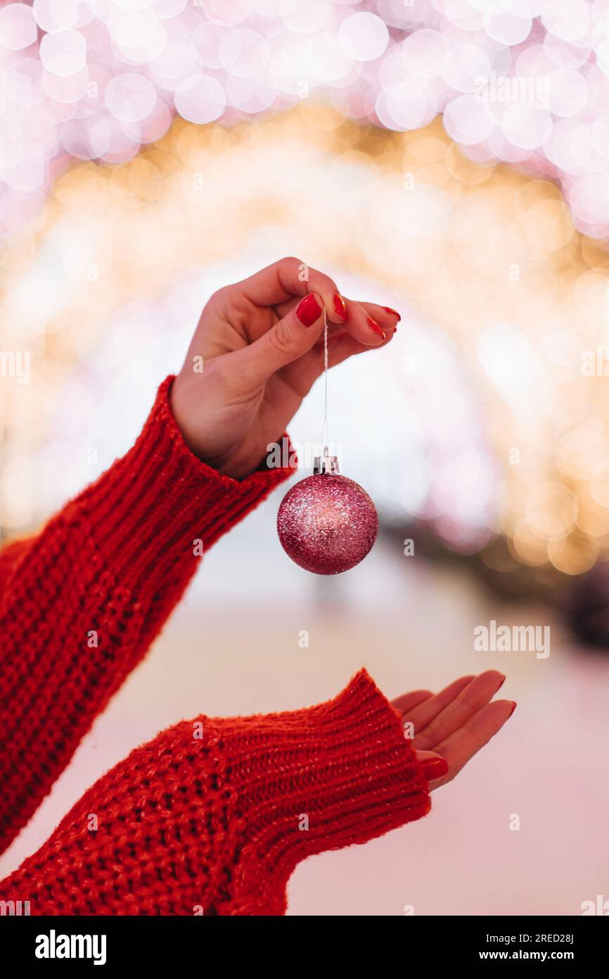 Weibliche Hände in einem gestrickten roten, gemütlichen Pullover mit einem pinkfarbenen Weihnachtsball vor einem festlichen goldenen Hintergrund Stockfoto