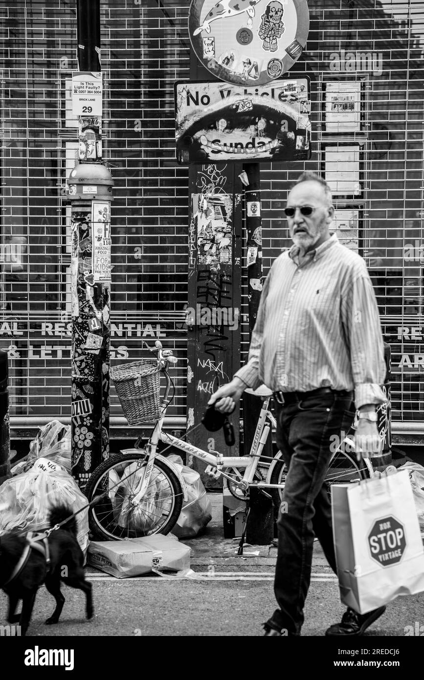 Männliche Figur mittleren Alters mit klassischer Einkaufstasche, die mit einem kleinen Hund in Brick Lane, London, England, Großbritannien spaziert Stockfoto