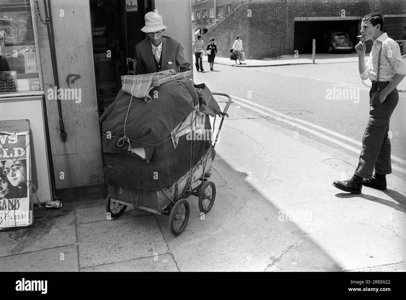 Skinhead oder Bovver Boy, der an einer Straßenecke rumhängt. Er trägt eine Hosenträger und kurze Hosen, damit man seine Bovver-Stiefel sehen kann, die gut waren, um jemandem einen „guten tritt“ zu geben. Der Look war modisch. Er schaut auf einen Obdachlosen, der all seine Besitztümer in einem Kinderwagen gestapelt hat. Tower Hamlets East London 1978. 1970S GB HOMER SYKES Stockfoto