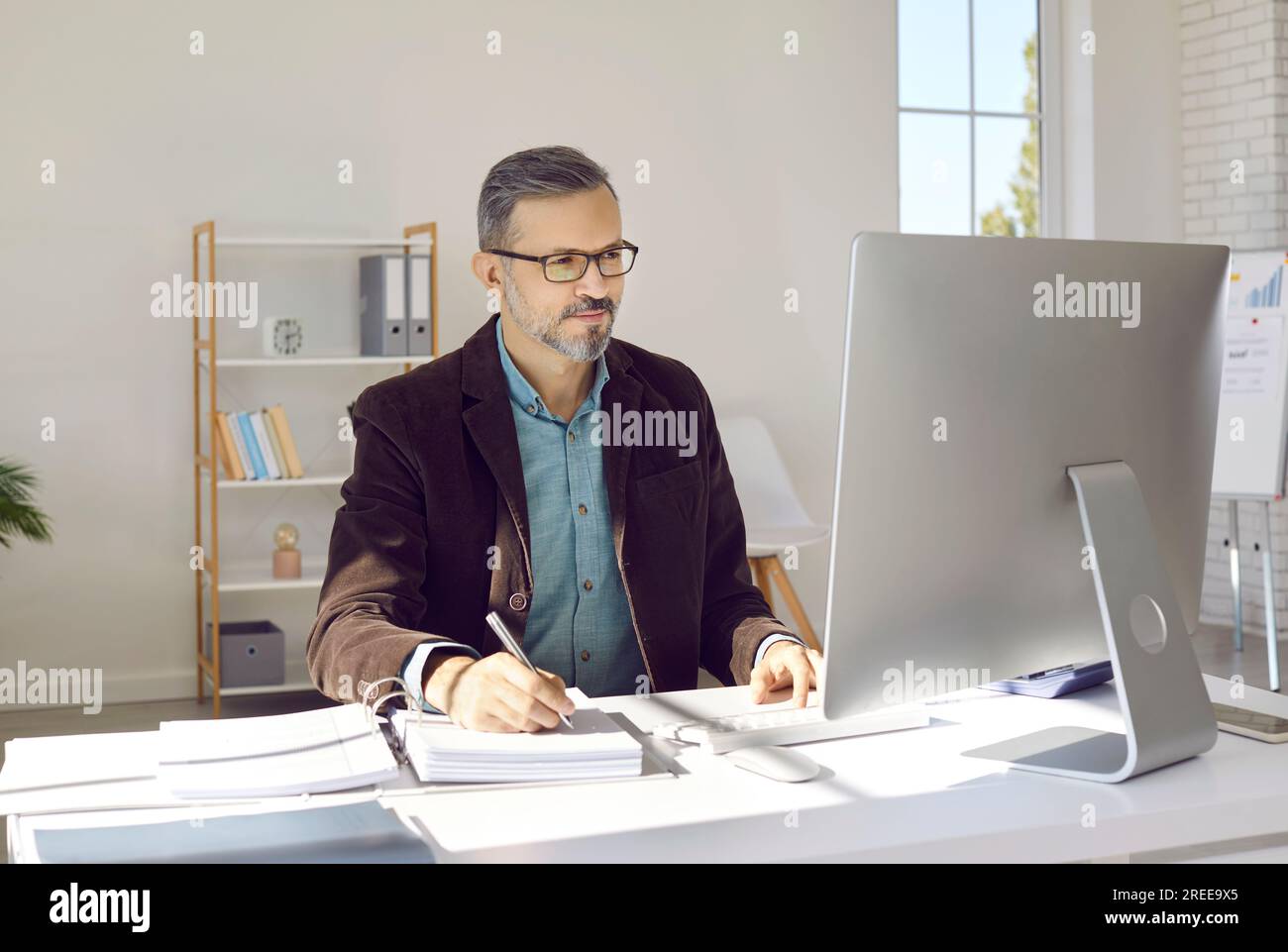 Porträt eines lächelnden, grauhaarigen Geschäftsmanns mit Brille, der vor seinem Computerbildschirm in einem modernen, hellen und geräumigen Büro sitzt. Stockfoto