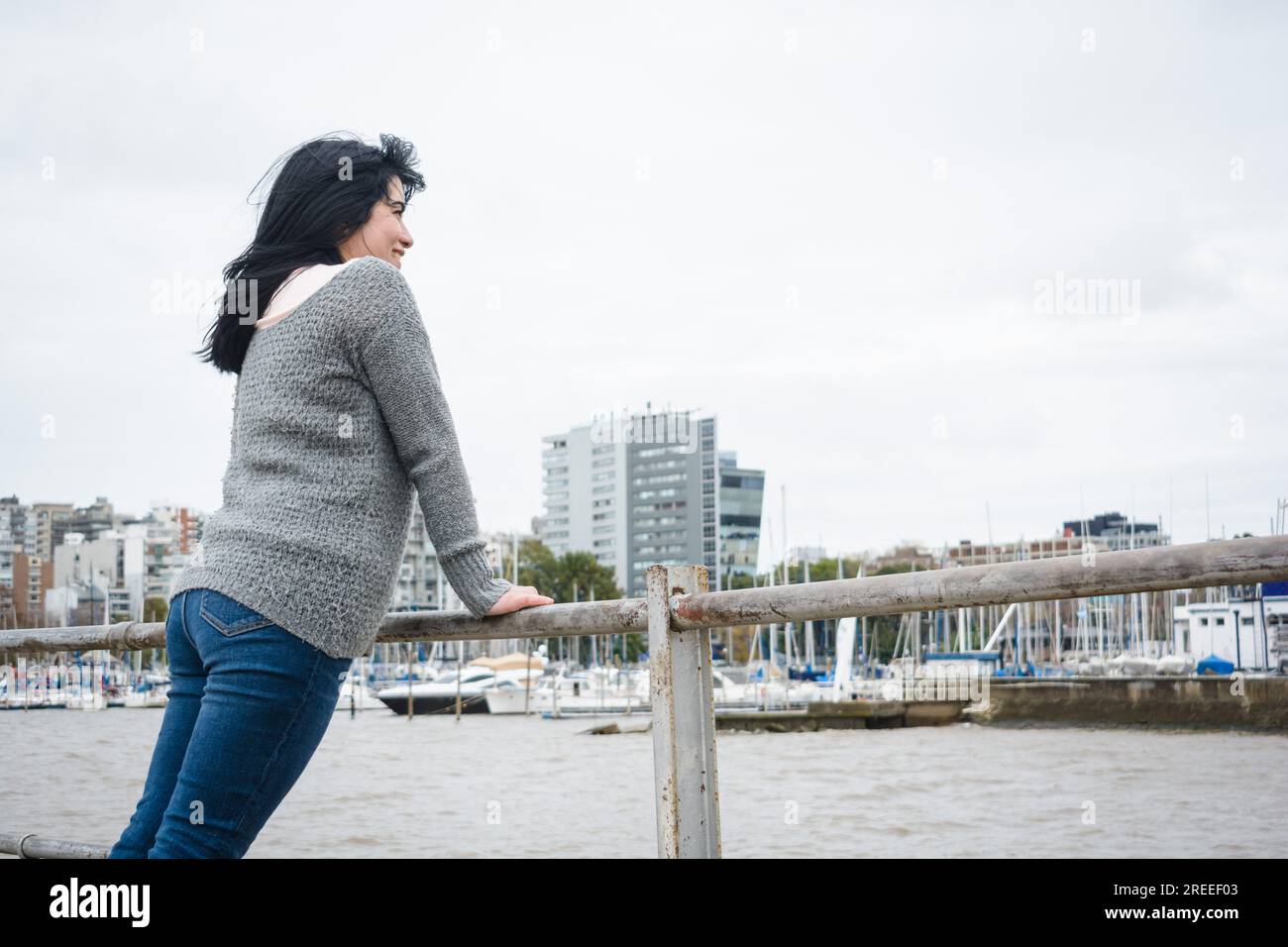 Junge abenteuerlustige Touristenlatina in einem grauen Pullover und einer blauen jeans-Hose, steht auf dem Geländer des Piers über dem Fluss und besucht Stockfoto