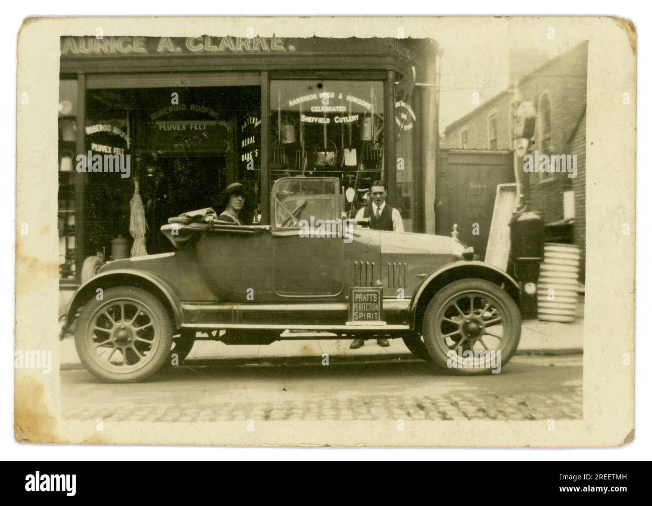 Original 1920 Jahre kleine Postkarte Maurice A. Clarke Ironmongers/Eisenwarenladen - kleines Familienunternehmen - die Frau des Eigentümers sitzt im Auto, Morris Cowley 'Bullnose', 2-Sitzer, möglicherweise zeigt sie das neue Familienauto. London, Großbritannien Ungefähr 1925. Stockfoto
