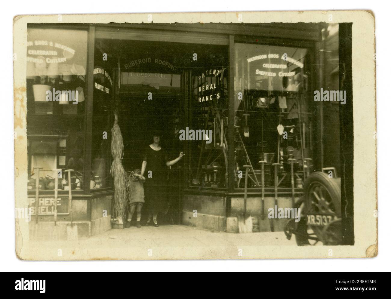 Maurice A. Clarke Ironmongers/Eisenwarenladen aus den 1920er Jahren – die Frau der Eigentümerin steht mit ihrem Kind in einer Tür. Vor dem Laden steht der Familienwagen Morris Cowley, die „Bullnose“, für 2 Personen geöffnet. Die Rückseite ist nur mit dem Buchstaben XR zu sehen, der auf eine Londoner Zulassung hinweist, Großbritannien Ungefähr 1925 Stockfoto