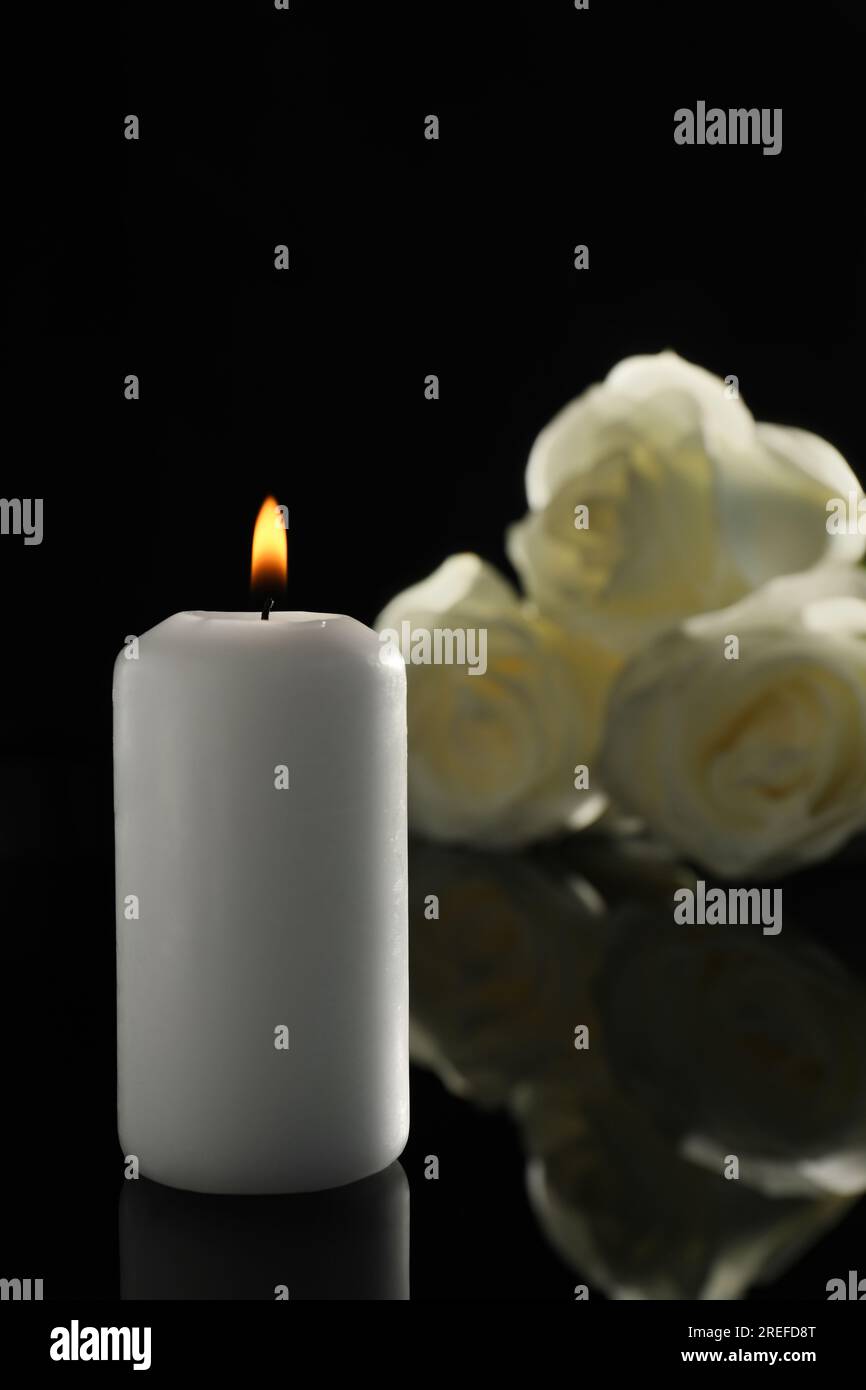 Weiße Rosen und brennende Kerze auf schwarzer Spiegeloberfläche bei Dunkelheit. Begräbnissymbole Stockfoto