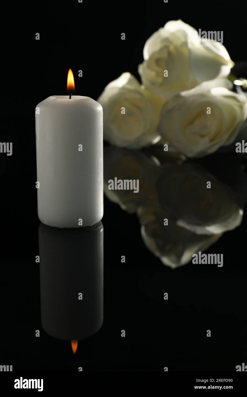 Weiße Rosen und brennende Kerze auf schwarzer Spiegeloberfläche bei Dunkelheit. Begräbnissymbole Stockfoto
