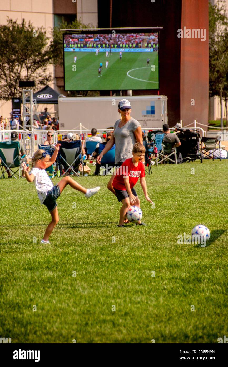 Costa Mesa, Kalifornien, USA. 15. Juli 2018. Während das Publikum tagsüber ein Fußballspiel der Weltmeisterschaft auf einem riesigen Fernsehbildschirm im Freien in Costa Mesa, CA, ansieht, spielt eine Mutter das Spiel mit ihren Kindern auf einem nahe gelegenen Spielfeld. (Kreditbild: © Spencer Grant/ZUMA Press Wire) NUR REDAKTIONELLE VERWENDUNG! Nicht für den kommerziellen GEBRAUCH! Stockfoto