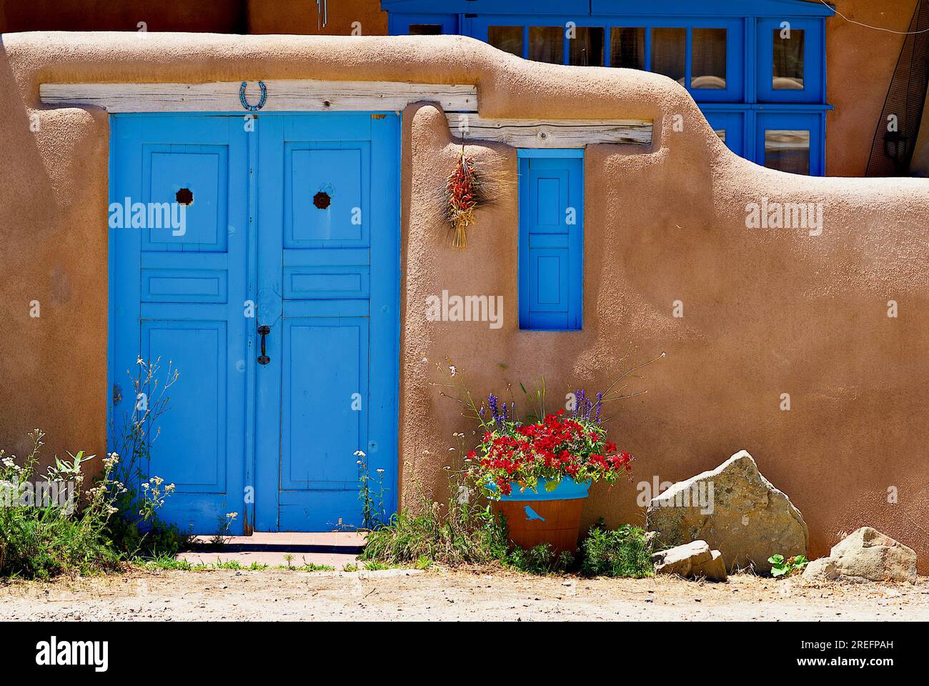 Rancho de Taos, New Mexico, USA - 24. Juli 2023: Eine blau gestrichene Tür und Rollläden im Kontrast zu den braunen lehmwänden eines Privathauses. Stockfoto