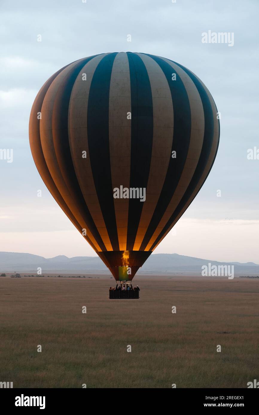 Blau-weiß gestreifter Heißluftballon mit Feuerlicht, Flug über die Savanne des Maasai/Masai Mara Reservats an einem bewölkten Tag, Berge am Horizont. Stockfoto