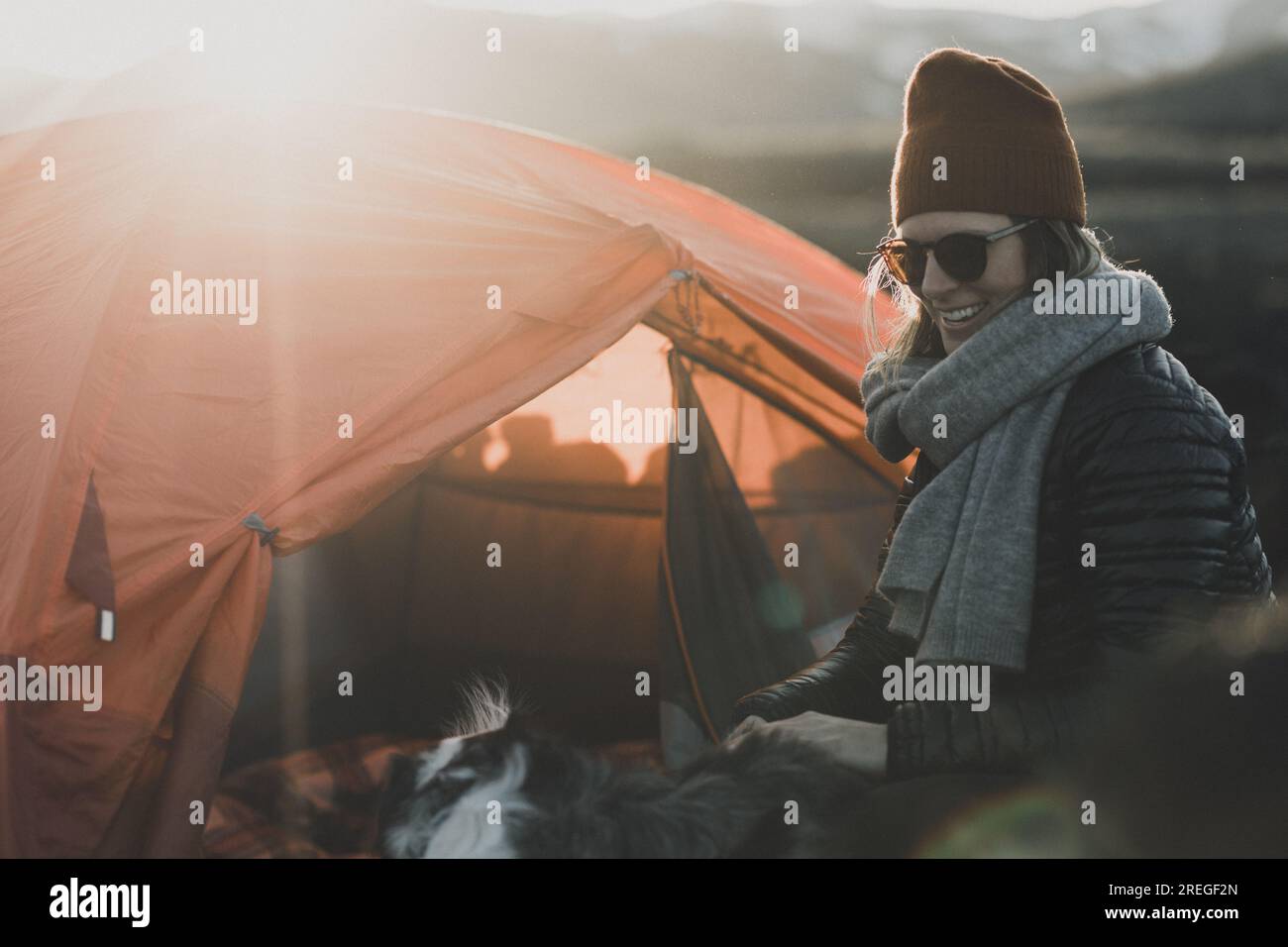 Fröhliche Campingfrau in Beanie bei Sonnenuntergang vor dem orangefarbenen Zelt Stockfoto