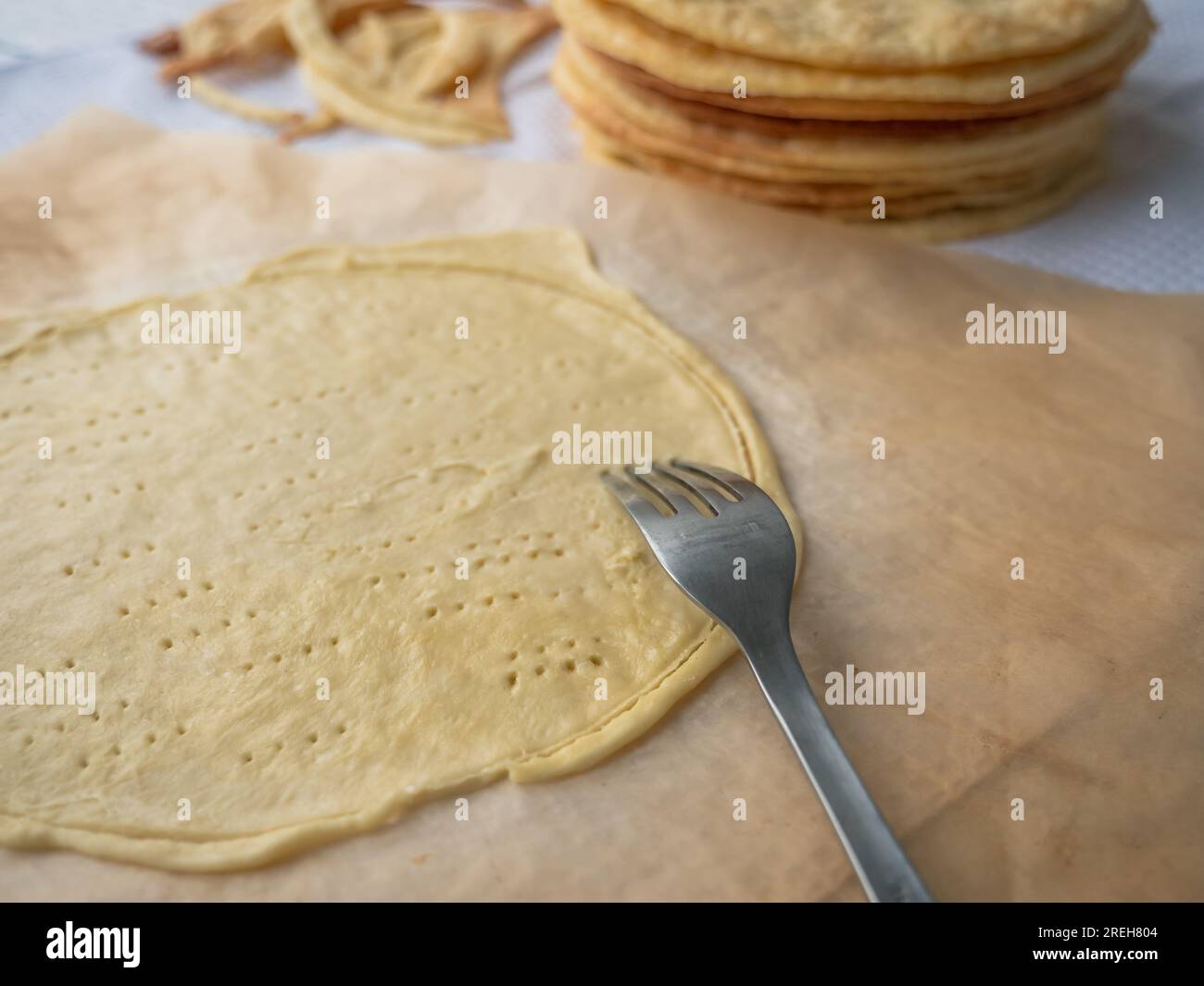 Mit der Gabel durchstechen Sie eine Rohschicht, die auf Backpapier ausgerollt ist, einen Haufen von gebackenen dünnen Butterschichten und Teigschrott im Hintergrund, um eine kurze Kruste zu bilden Stockfoto