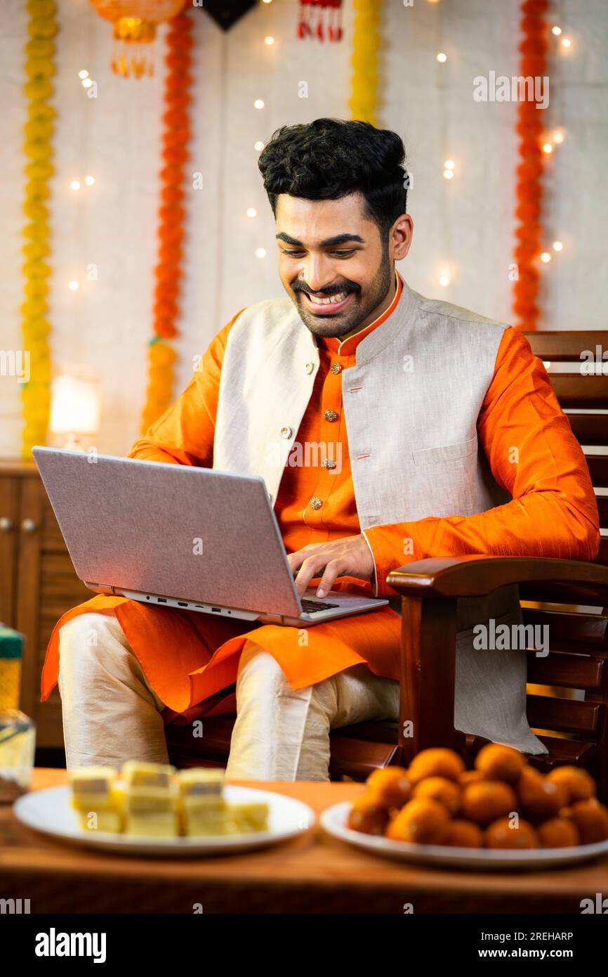 Glücklicher junger indischer Mann, der während der diwali Festival Feier im dekorierten Haus ein Notebook benutzte, vor Geschenken und Geschenken, indem er in die Kamera schaute - Konzept Stockfoto