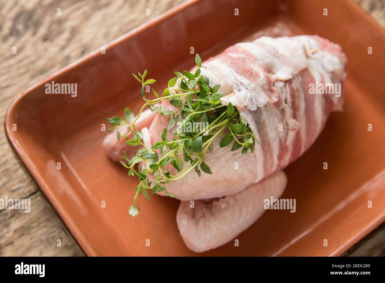 Ein rohes, ungekochtes Hühnerfilet auf dem Knochenviertel, das in durchwachsenen Speck eingewickelt und mit frischem Thymian aromatisiert wurde. Es wird in Weiß geschmort Stockfoto