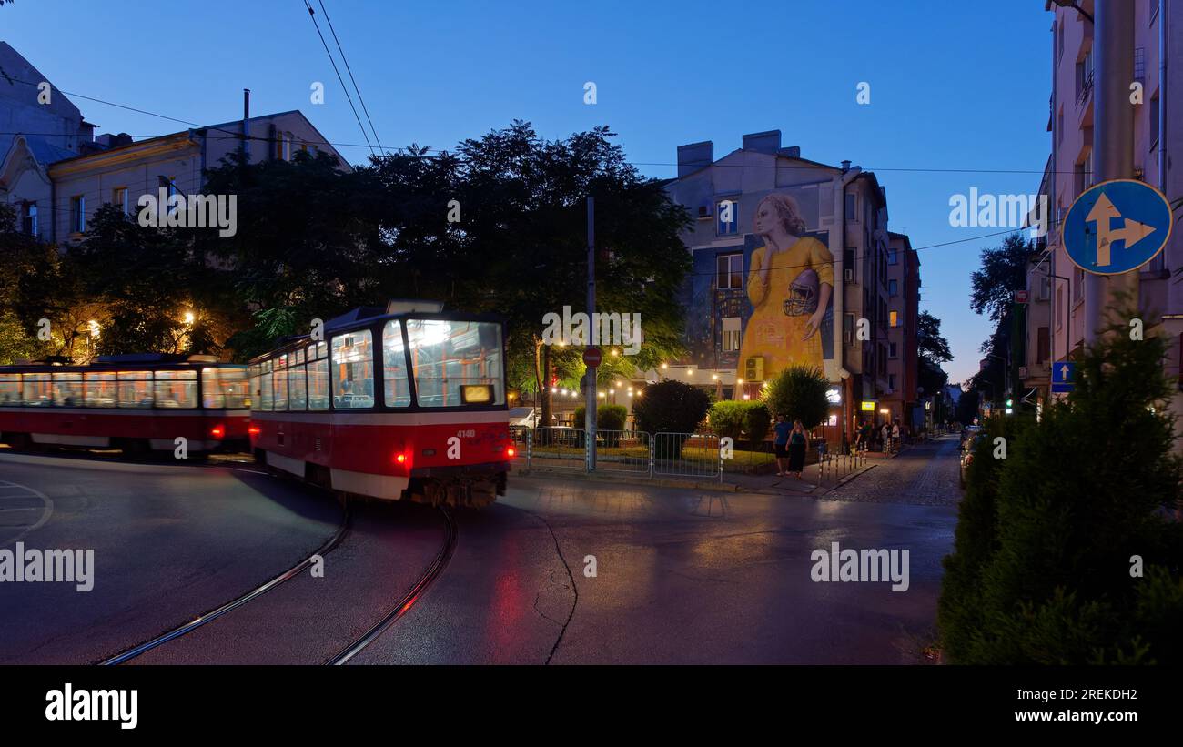 Die Straßenbahn leuchtet nachts in Sofia, Bulgarien, mit Straßenkunst einer Dame in einem gelben Kleid und einem Helm unter dem Arm. Sofia, Bulgarien. Juli 2023 Stockfoto