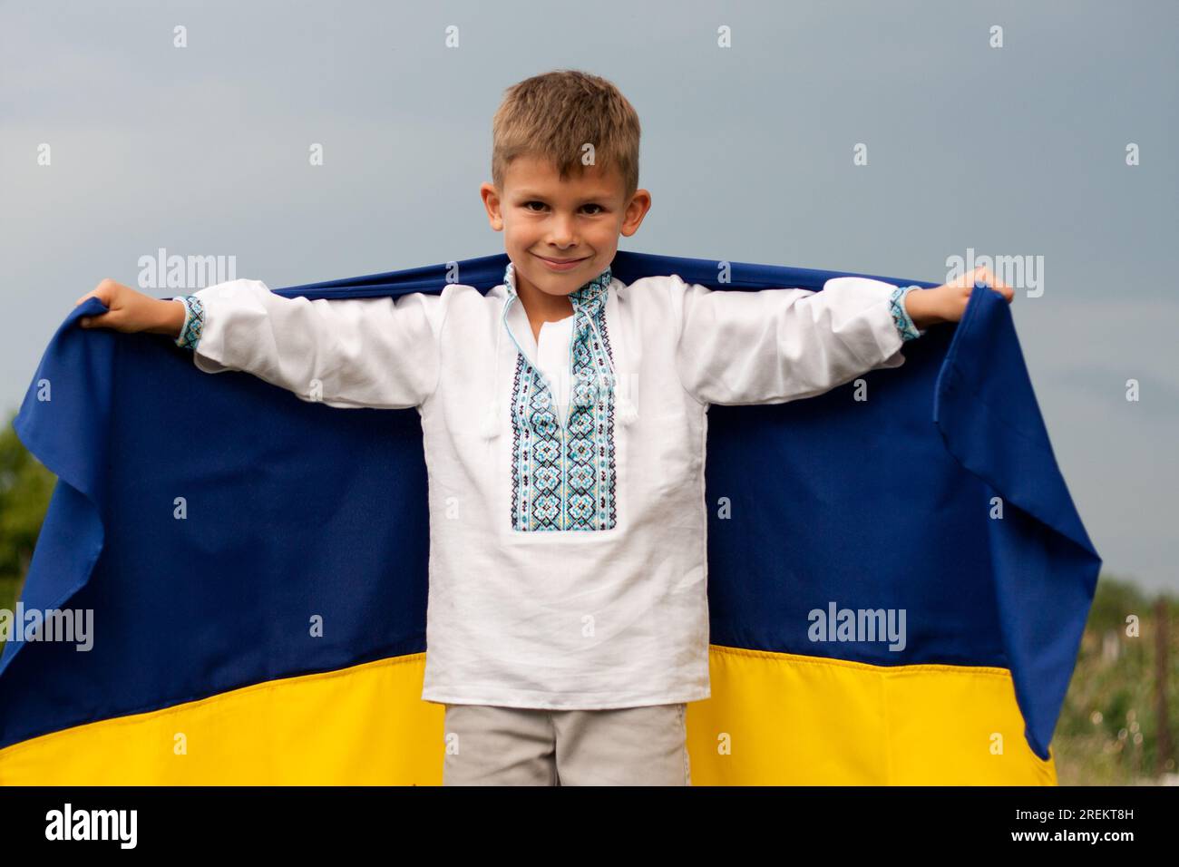 Glücklicher ukrainischer Junge in besticktem Hemd - vyshyvanka mit blau-gelber Flagge der Ukraine, gegen den Himmel. Unabhängigkeitstag der Ukraine. Verfassungstag. Da Stockfoto