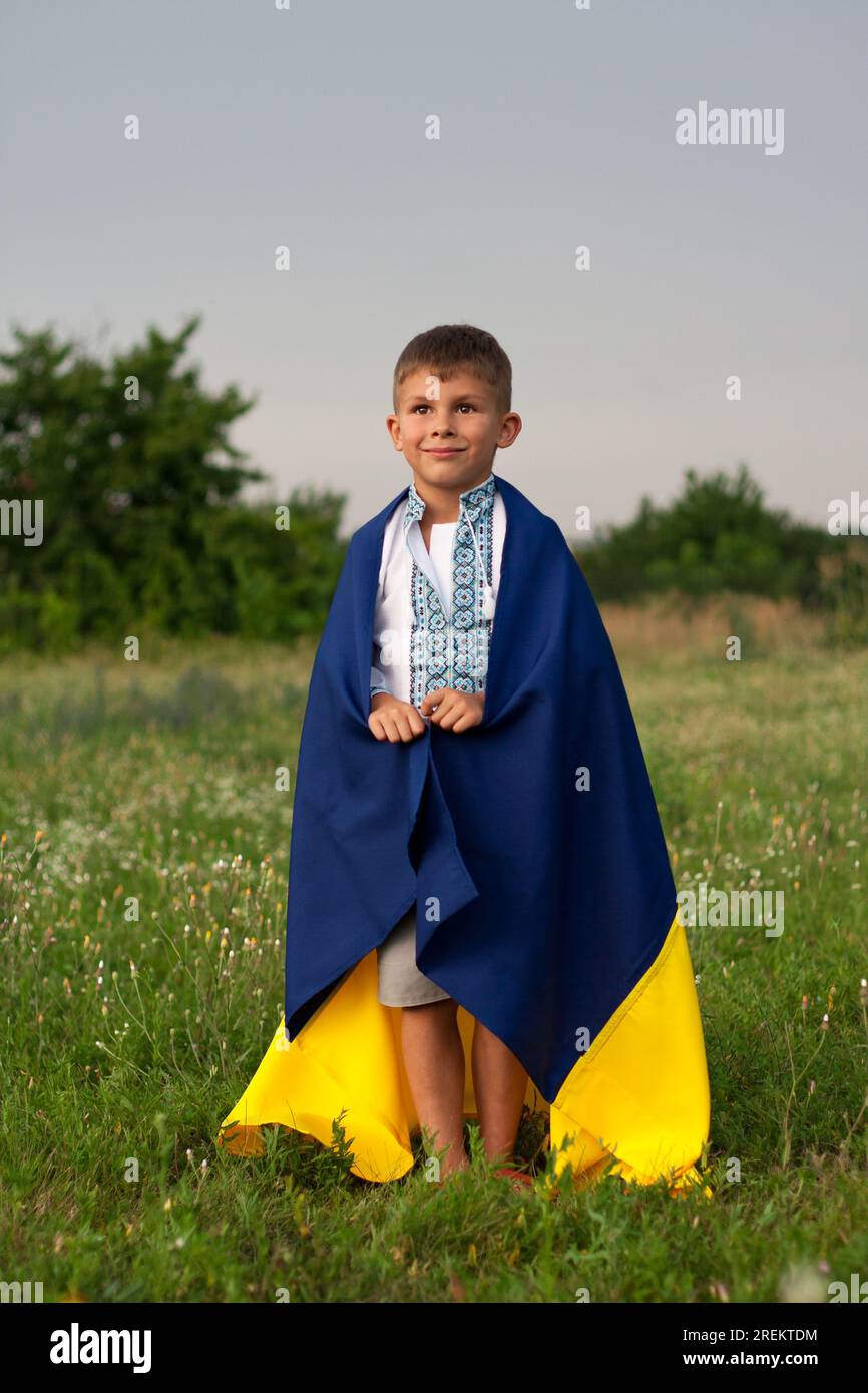 Glücklicher ukrainischer Junge in besticktem Hemd - vyshyvanka mit blau-gelber Flagge der Ukraine, auf Wiese am Sommertag. Unabhängigkeitstag der Ukraine. Constitu Stockfoto