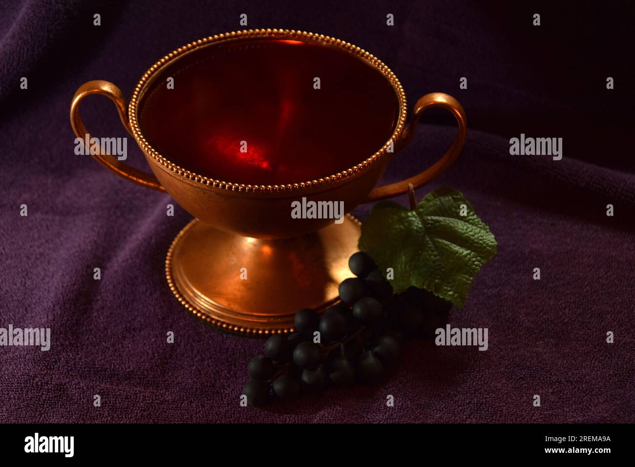 Goldfarbener Kelch mit roter Flüssigkeit und Trauben daneben auf einem violetten Tuch Stockfoto