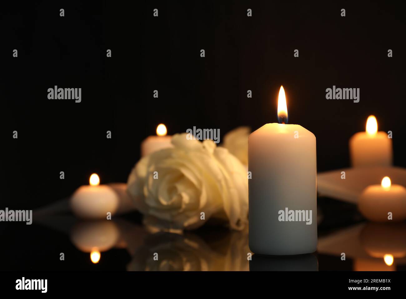 Weiße Rose und brennende Kerzen auf schwarzer Spiegeloberfläche bei Dunkelheit, Nahaufnahme mit Platz für Text. Begräbnissymbole Stockfoto