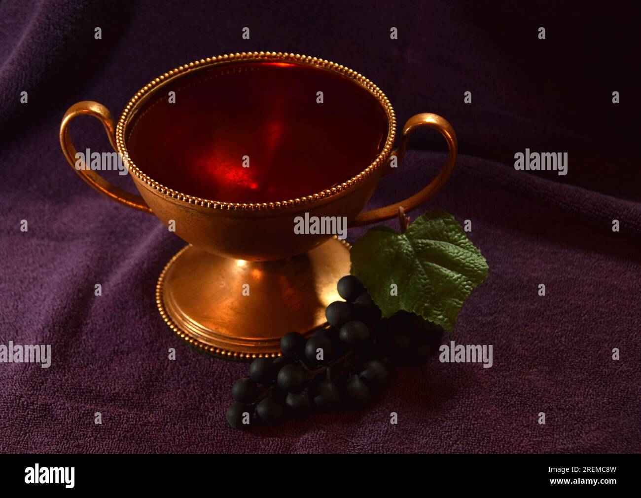 Goldfarbener Kelch mit roter Flüssigkeit und Trauben daneben auf einem violetten Tuch Stockfoto