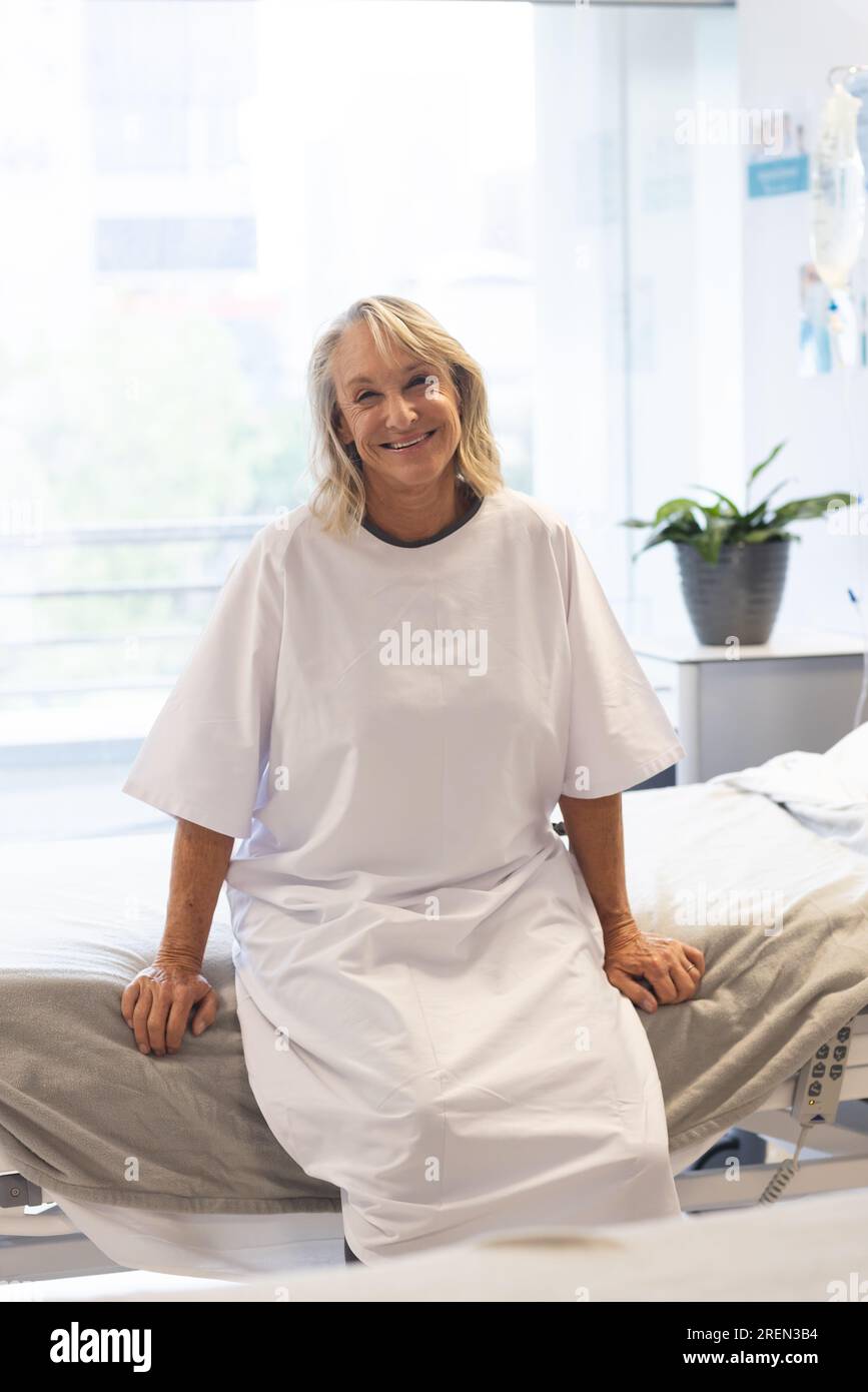 Portrait der glücklichen älteren weissen Patientin trägt Krankenhauskittel sitzend auf dem Bett im Krankenhaus Stockfoto