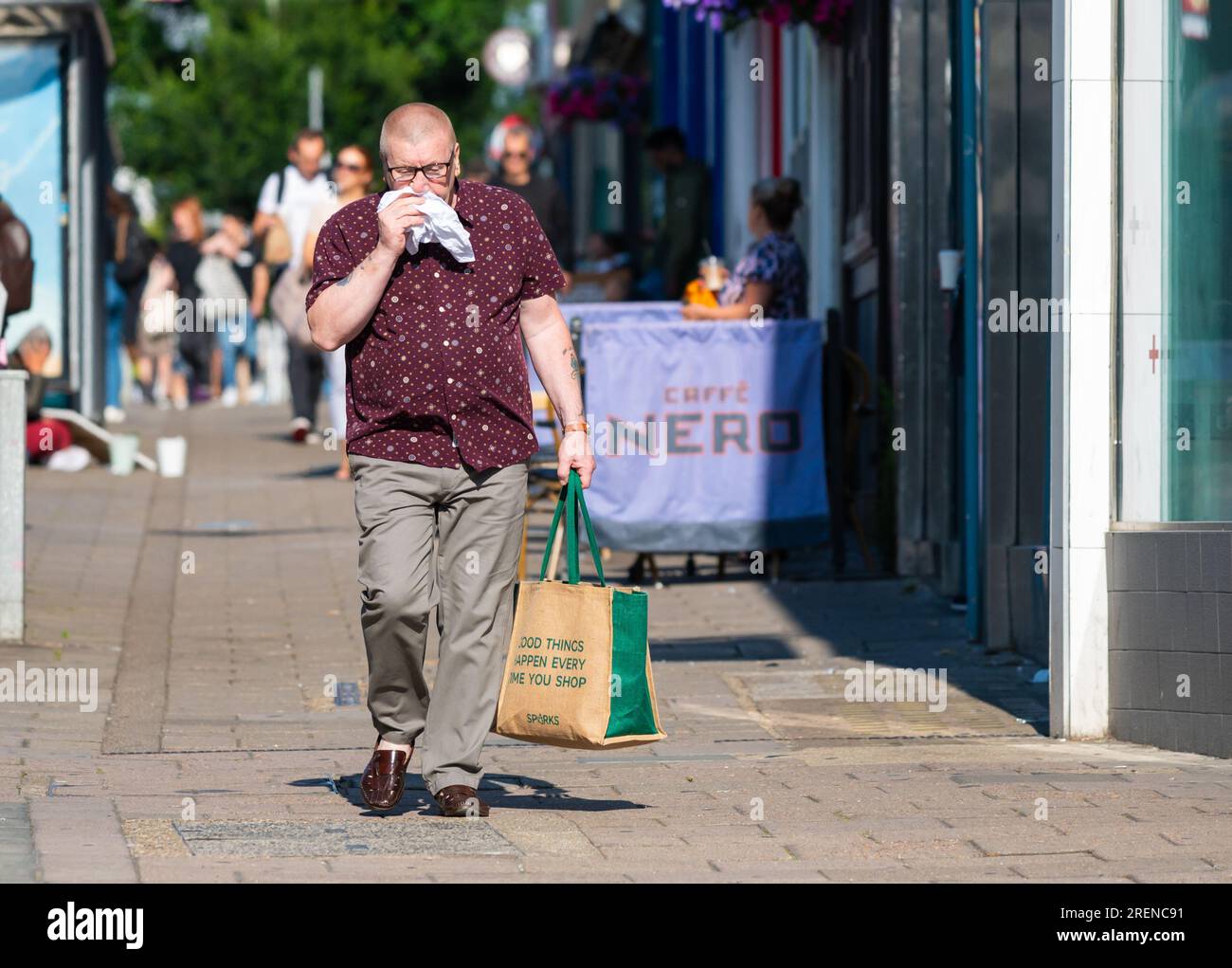 Ein Mann geht, schlau gekleidet in geknöpften Hemden und Hosen, benutzt Taschentuch oder Taschentuch, um die Nase zu putzen, möglicherweise Heuschnupfen oder eine Erkältung, Summer in Großbritannien. Stockfoto