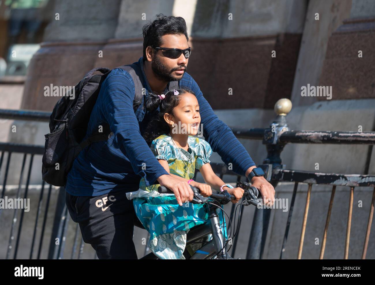 Mann und kleines Kind, die zusammen auf einem Fahrrad auf einer Hauptstraße radeln, ohne Helme zu tragen. Radfahrer ohne Helm in Großbritannien. Kindersicherheit. Stockfoto